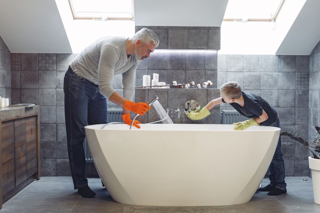 Side view of grey haired father with beard in orange rubber gloves and small kid cleaning bathroom together
